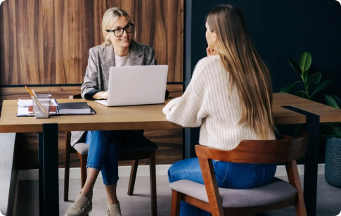 Two women sit at a wooden table talking, one of them is using a laptop computer.