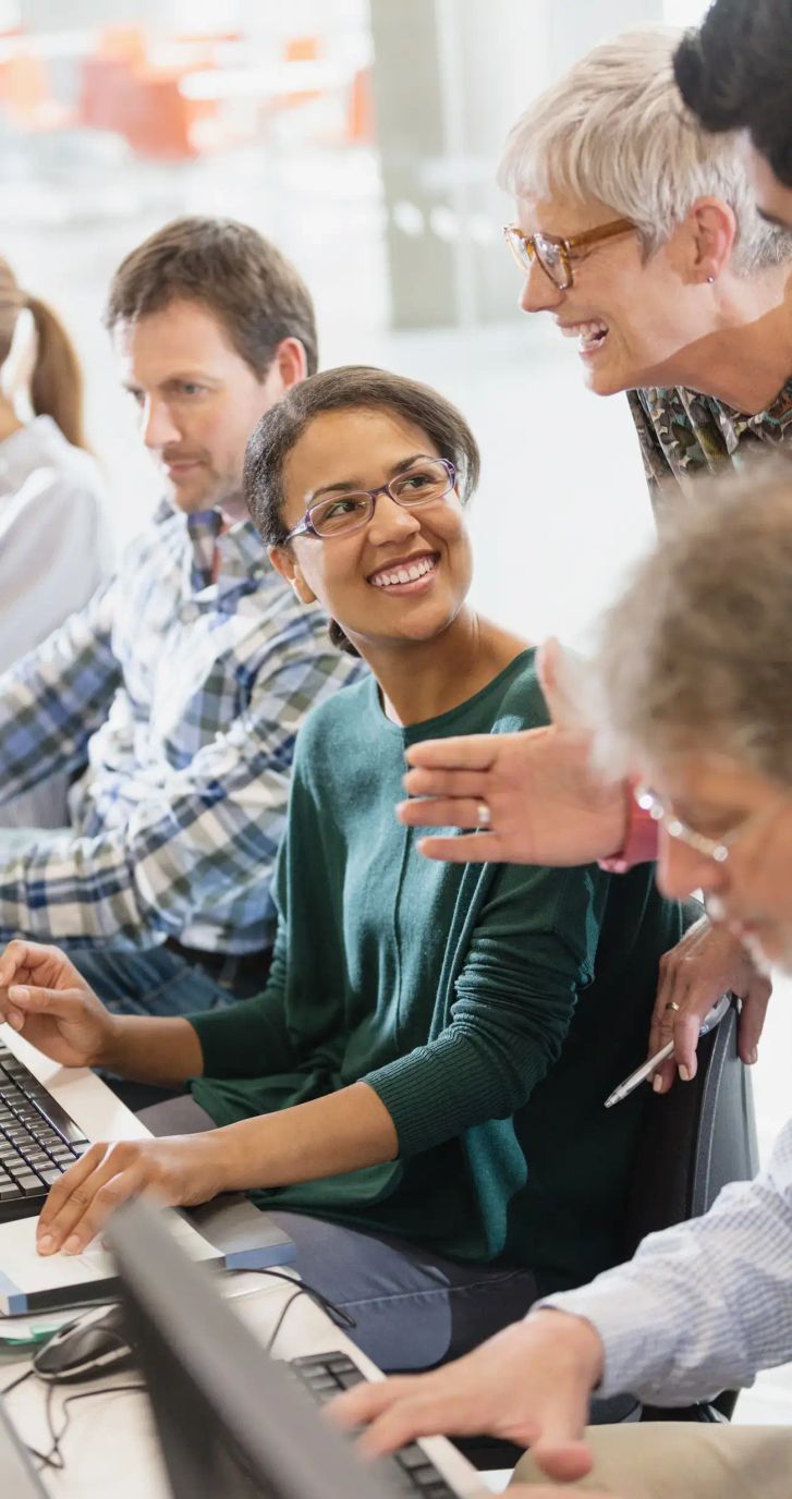 A group of people sitting at computers with a lady standing behind them.