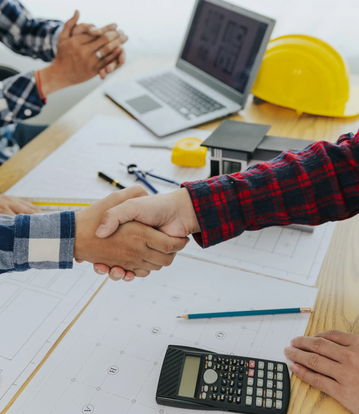 Two people shake hands over a desk covered in paperwork.