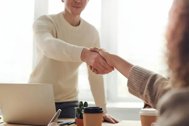 Two people shaking hands across a desk with a coffee cup and pens.