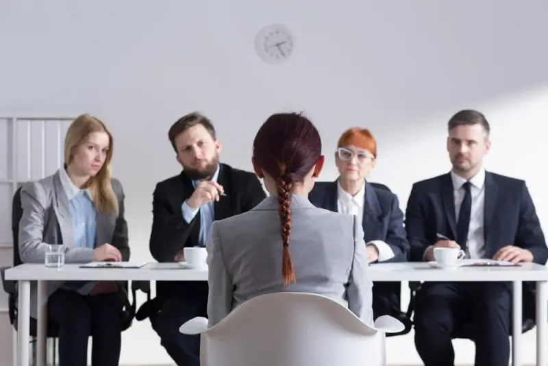 Four people conduct an interview from behind a desk while the lone interviewee sits in front of them on a chair