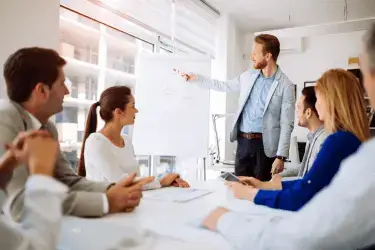 A group of people sit around a table while one of them is standing and pointing at a flip chart.