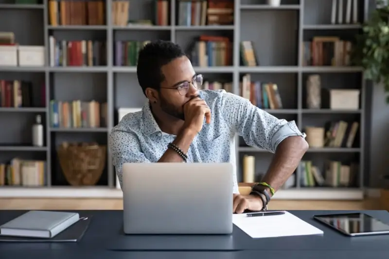 A man sits at his desk in front of his laptop thoughtfully looking out of the window.