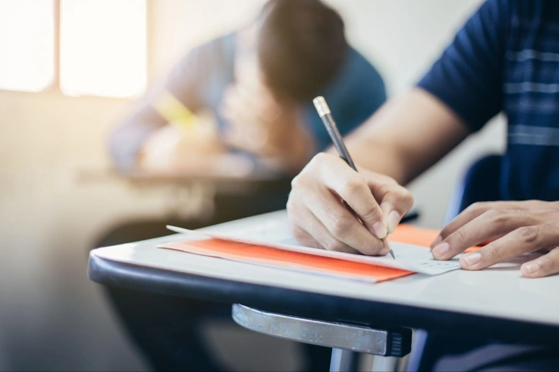 A close up as someone sits at a desk writing on a piece of paper with a pencil.