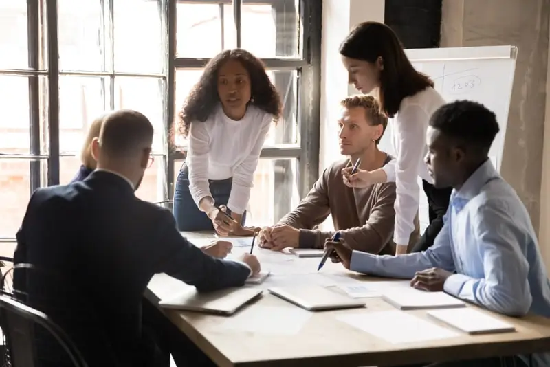 A group of people gather around a table taking notes as one of them speaks